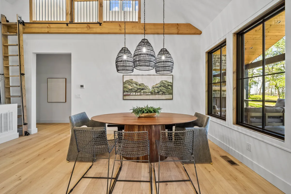 A sophisticated dining area with a large wooden table beneath a statement chandelier, surrounded by artwork and expansive windows that bathe the space in light. The scene suggests a space designed for shared meals and celebrations.