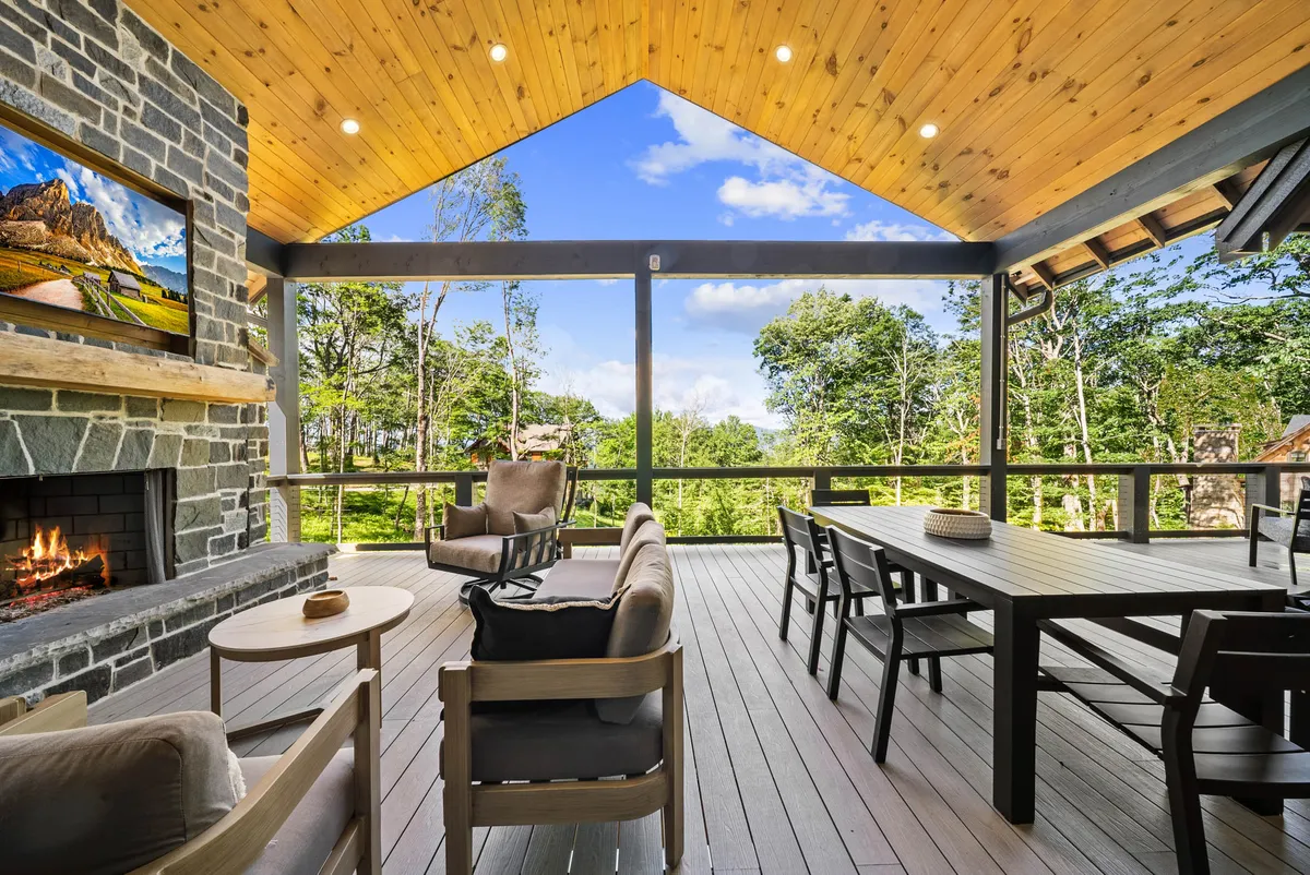 A spacious exterior deck features a striking stone fireplace as its centerpiece, surrounded by plush outdoor seating and a large dining table. The deck provides sweeping views of the surrounding natural landscape, and large windows connect the indoor living space to the outdoors, creating a seamless flow. The cozy furnishings and inviting atmosphere suggest it is perfect for both quiet evenings and lively gatherings.