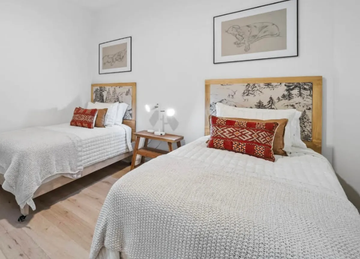A welcoming bedroom with two beds, decorative headboards, colorful throw pillows, a small bedside table with lamp, and framed artwork. Soft morning light filters into the room, creating a cozy and inviting environment ideal for families or extra guests.