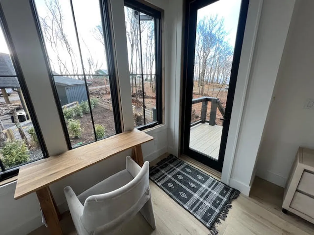 A cozy workspace setup features a comfortable chair and wooden table positioned by large windows, with a door leading outside. The room is accented by a textured rug, creating a calm and productive environment perfect for work or relaxation while enjoying views of nature.