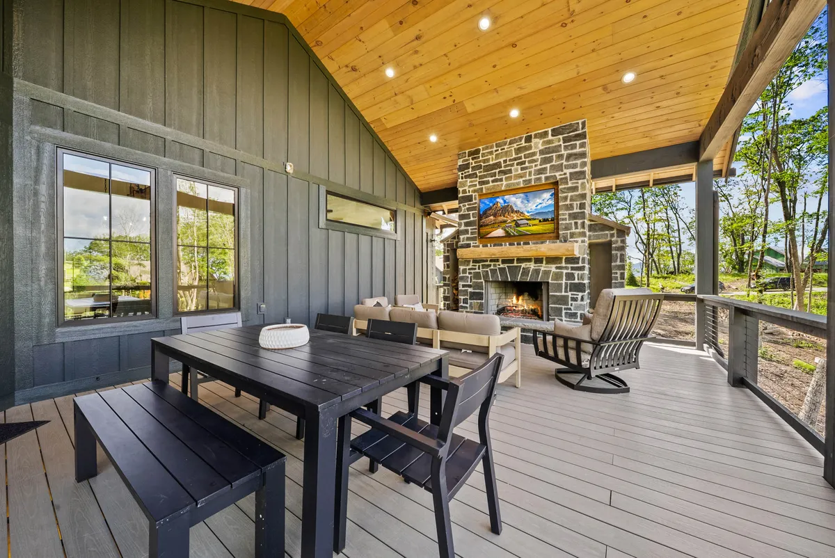 An inviting covered outdoor area featuring a stone fireplace, a generous dining table, comfortable seating, and large windows looking into the house. The setup is ideal for group dining, socializing, and relaxing by the fire, with space for everyone to gather in comfort.