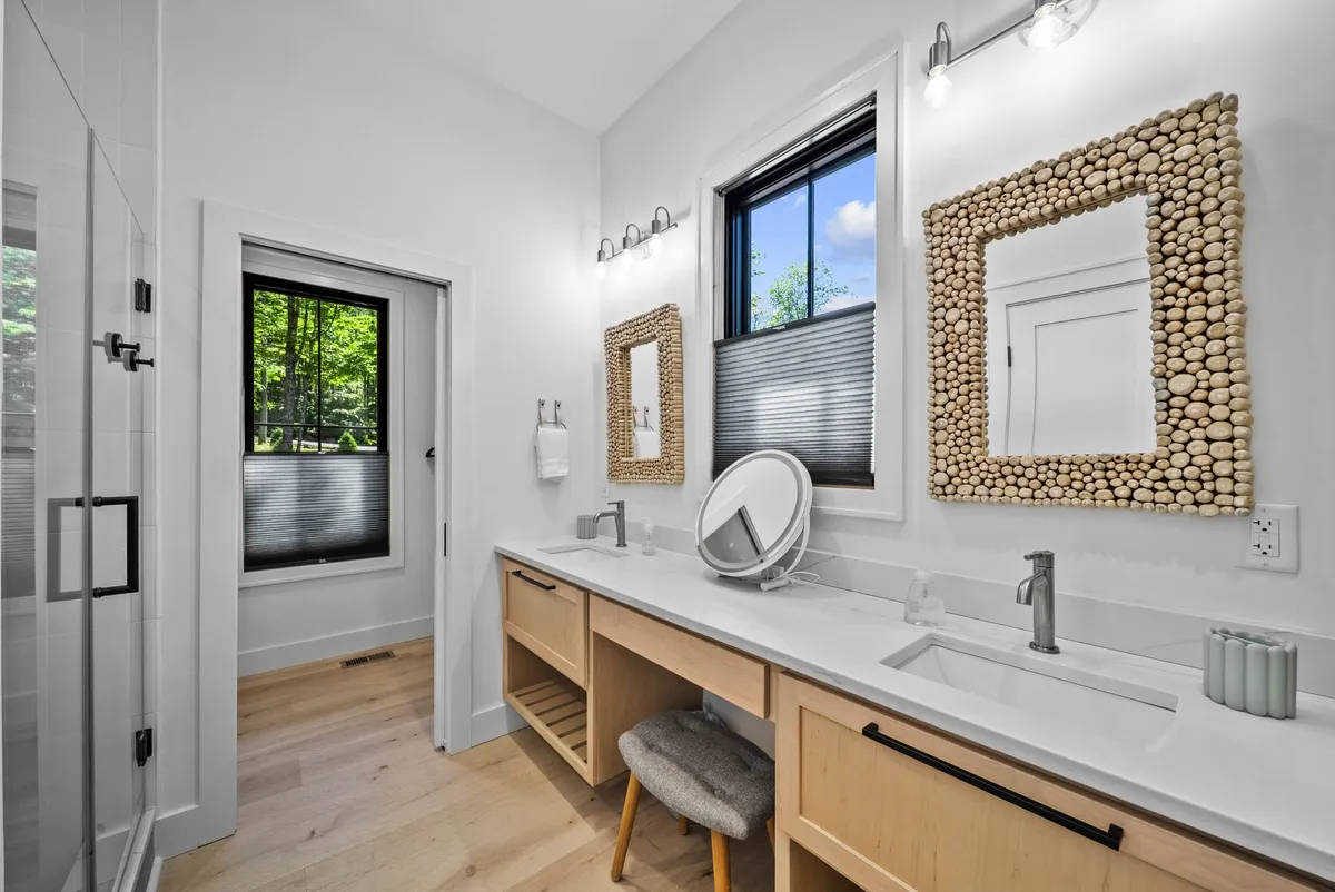 A modern ensuite bathroom with a double vanity featuring dual sinks and decorative mirrors. Natural light pours in from a large window, and a sleek shower stall is visible, exuding a spa-like ambiance ideal for self-care routines.