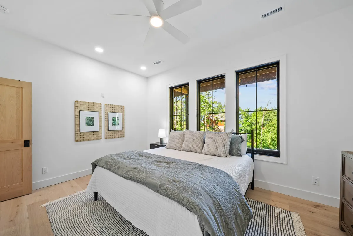 This image shows a tastefully decorated bedroom with a king-size bed adorned with decorative pillows. Large windows flood the room with natural light, and artwork on the wall adds a personal touch. The tranquil mood and ample sunlight create a restful, sophisticated ambiance.