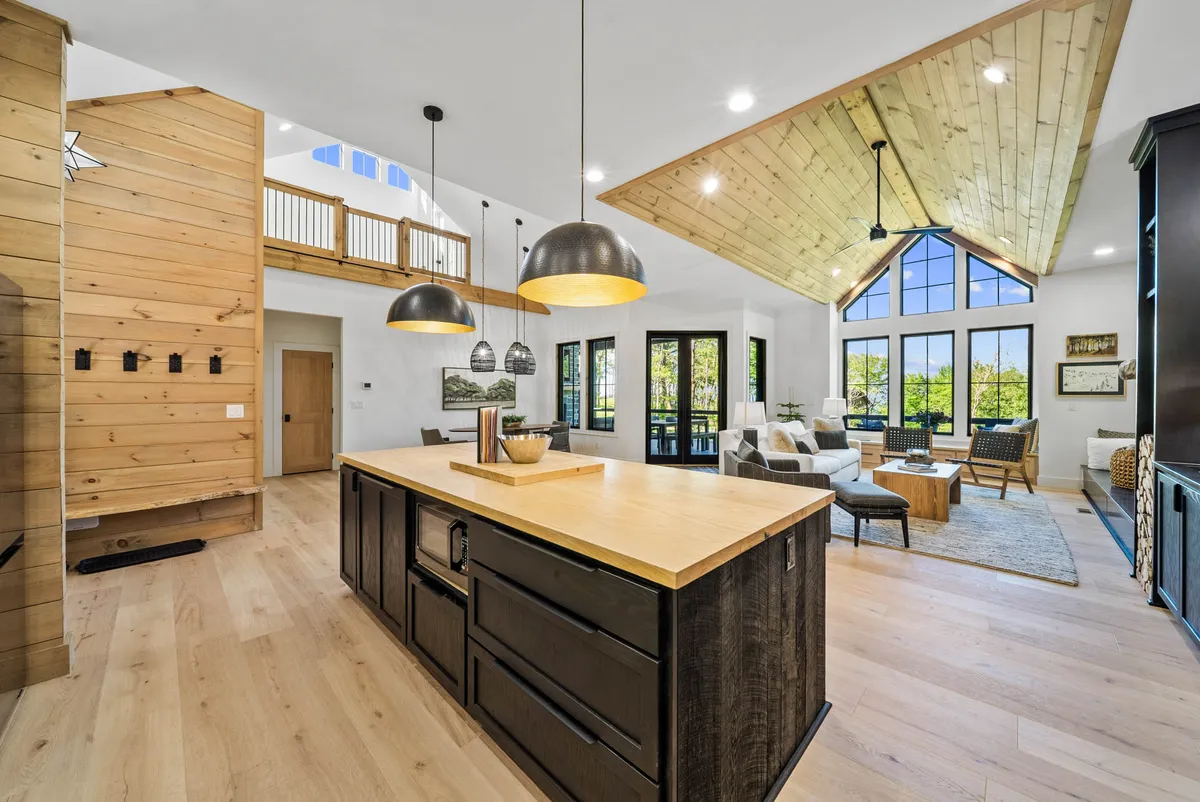 The image showcases an expansive, sunlit kitchen and great room area featuring a large kitchen island, vaulted ceiling with rustic wooden beams, and large windows that flood the space with natural light. Comfortable seating is arranged near the kitchen, creating a warm, open atmosphere ideal for gathering and entertaining.