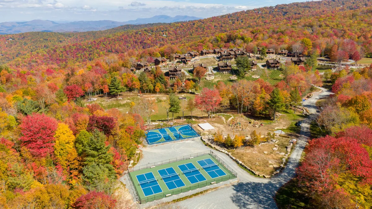 This image captures tennis courts nestled among colorful autumn trees, with a scenic mountain backdrop and residential buildings nearby. The courts are clearly visible and look well maintained, suggesting active use for sports and recreation. The surrounding mountain landscape hints at the broader outdoor opportunities available in the area.