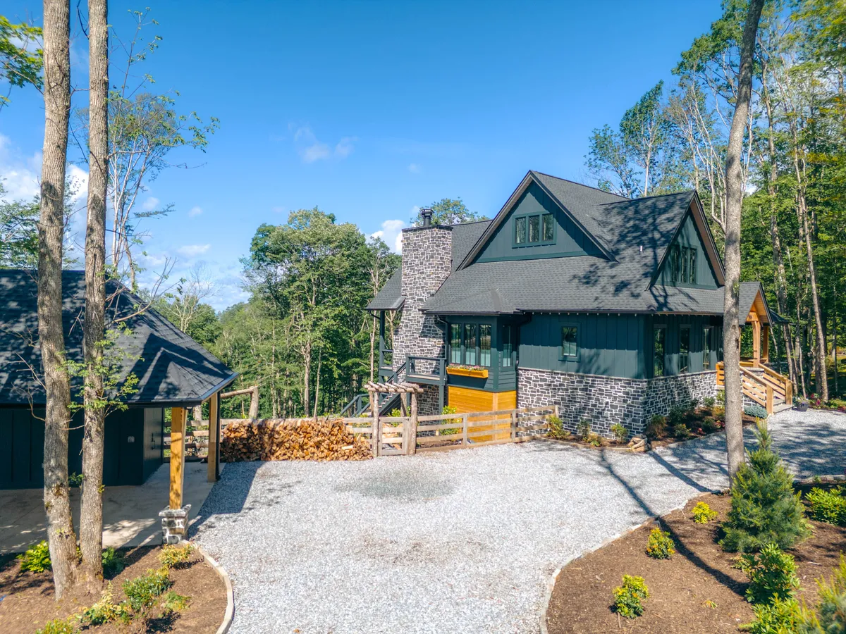 The hero image showcases the charming exterior of Lost Bark Lodge—a rustic yet inviting cabin nestled among tall trees in Banner Elk. The stone chimney hints at cozy nights by the fireplace, while a welcoming path leads to the main entrance. Visible are inviting outdoor spaces, perfect for gathering around the fire pit, soaking up mountain sunsets from the porch, or stargazing under clear Carolina skies.