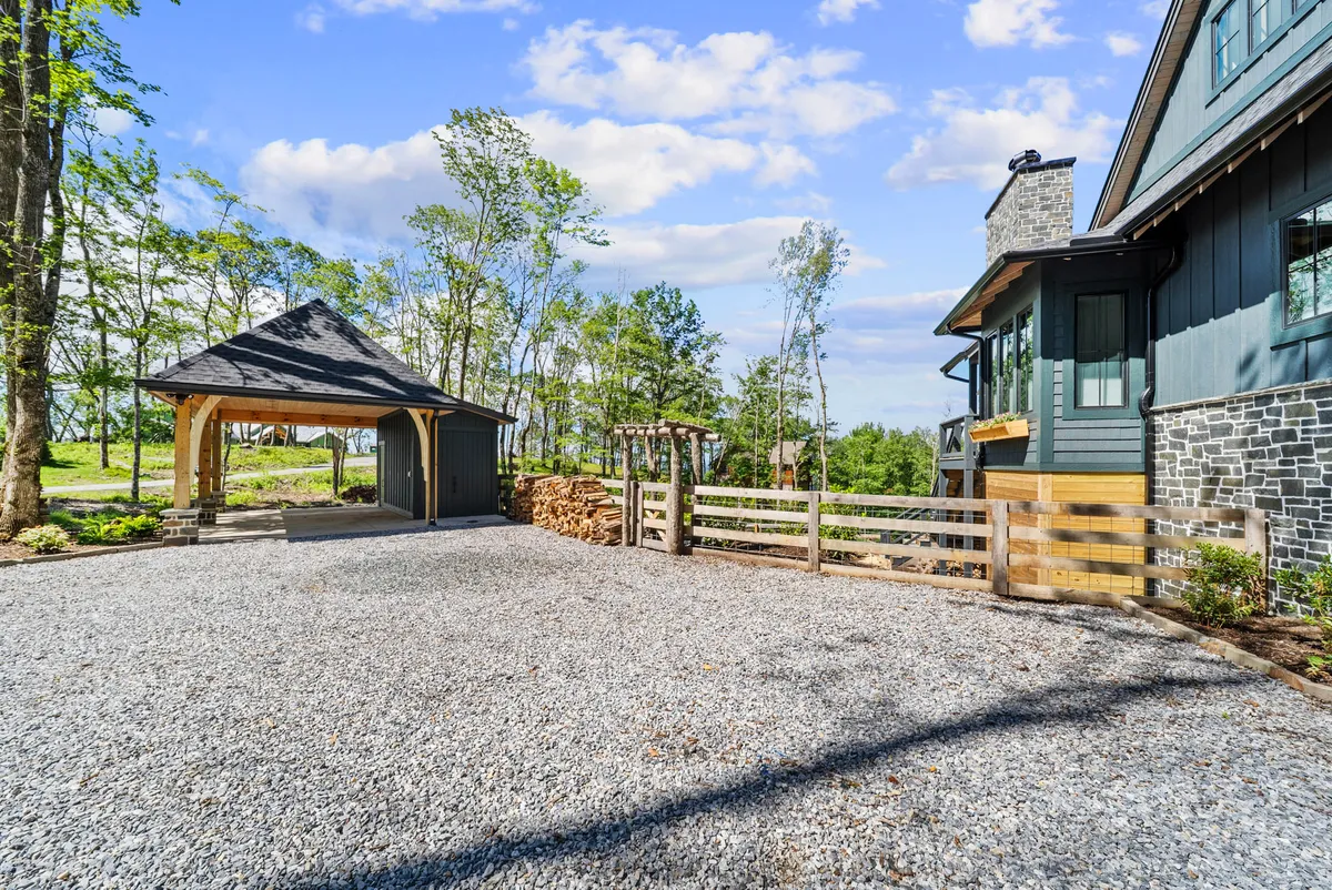 This image features a welcoming exterior shot of Lost Bark Lodge, showing the stone house, a wooden fence, and a spacious gazebo within a fenced yard. The gravel driveway offers a sense of openness, and the wooden fence is prominent, clearly defining a secure area for pets to play. The landscaped garden adds a homey, nature-oriented feel, perfect for those traveling with dogs.